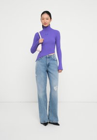 Maglione a collo alto viola a coste con maniche lunghe, abbinato a jeans chiari a gamba larga e strappati e scarpe nere a punta. Piccola borsa rosa.