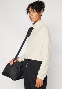 Pullover in pile beige chiaro con colletto alto, caratterizzato da una zip frontale a metà, indossato con pantaloni neri. Borsa nera a tracolla.