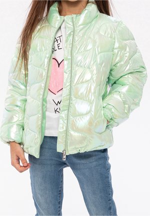 Enfant portant une doudoune vert clair brillante sur un t-shirt blanc avec un motif cœur rose et un jean bleu, debout avec une main dans la poche.