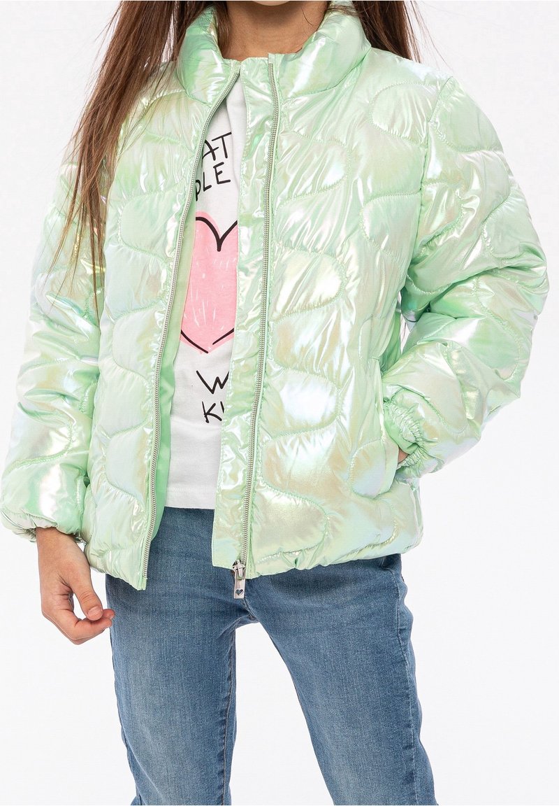 Enfant portant une doudoune vert clair brillante sur un t-shirt blanc avec un motif cœur rose et un jean bleu, debout avec une main dans la poche.