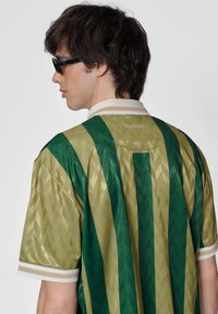 Grøn-og-guld-stribet sportstrøje med geometrisk mønster, der har en hvid krave og diskret logo placering på ryggen.
