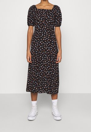 Person iført en sort midi kjole med pufærmer og små røde, blå og hvide blomsterprint, kombineret med hvide høje sneakers og sokker.