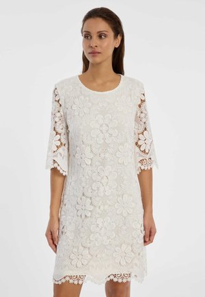 Vrouw met een witte bloemenkantjurk met halve mouwen en geschulpte randen, staand tegen een effen witte achtergrond.