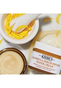 Kiehl's Calendula Serum-Infused Water Cream i en krukke med en hvit etikett, omringet av gule ringblomsterblader og en hvit morter og støter.