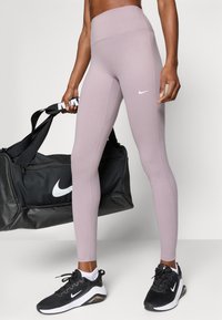 Leggings violet clair en tissu extensible, taille haute, avec un logo Nike blanc. Sac de sport noir avec logo blanc et bretelles.