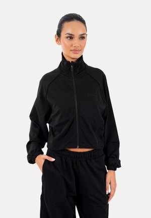 Femme aux cheveux foncés portant une veste noire zippée et un pantalon noir assorti à taille élastique, debout avec une main dans la poche.