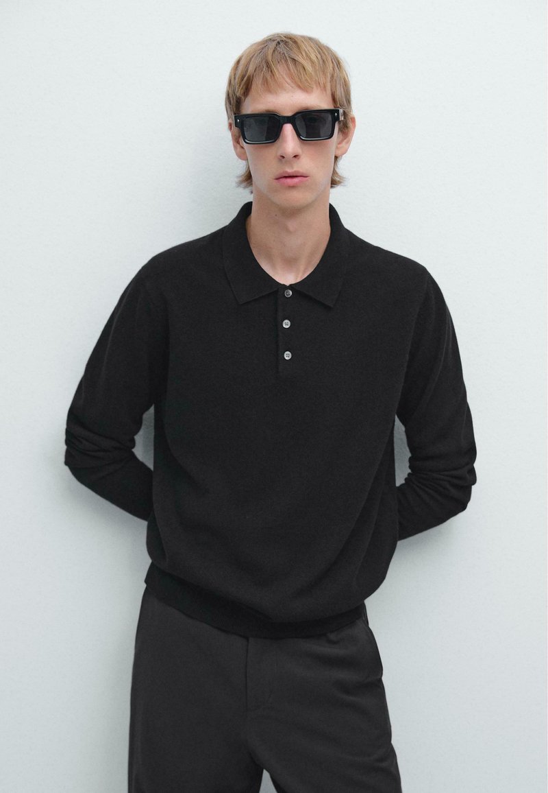 Schwarzes, gestricktes Polo mit langen Ärmeln, einem Kragen und drei Knöpfen. Getragen mit schwarzen Sonnenbrillen. Einfaches, tailliertes Design vor einem neutralen Hintergrund.
