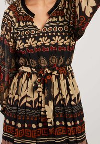 Femme portant une robe noire à manches longues avec des motifs géométriques et floraux beiges et roux, ceinture nouée à la taille et décolleté en V.