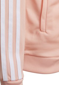 Rosa sweatshirttyg med ribbat mönster, med tre vita ränder längs ärmen. Inkluderar en framficka och elastiska mudd.