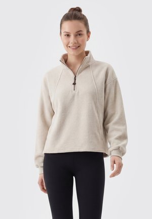 Ung kvinde i beige kvart-zip trøje og sorte leggings smilende mod en lys, ensfarvet baggrund.