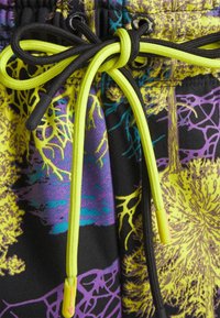 Close-up of neon yellow and black drawstrings tied in a bow on multicolored fabric with abstract yellow, purple, and blue patterns.