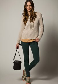 Donna con lunghi capelli castani, che indossa un maglione beige, pantaloni verdi e scarpe dorate, tiene in mano una piccola borsa nera su uno sfondo neutro.