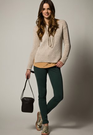 Vrouw met lang bruin haar, gekleed in een beige trui, groene broek en gouden schoenen, met een kleine zwarte handtas tegen een neutrale achtergrond.