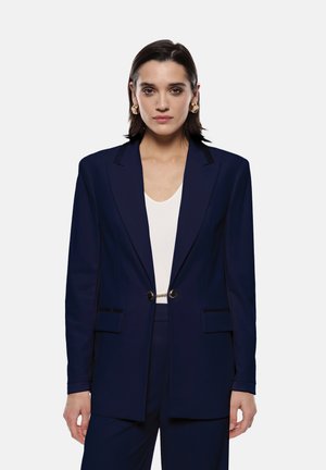 Blazer su misura di un profondo blu navy con rever a punta, due tasche frontali e chiusura a doppio bottone. Presenta un tessuto liscio e una vestibilità strutturata.