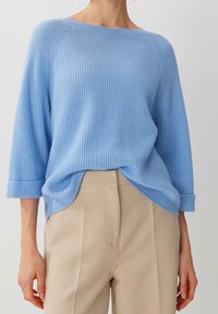 Pull léger bleu à côtes avec des manches trois-quarts, offrant une coupe décontractée et de légentes fentes sur les côtés à l'ourlet, associé à un pantalon beige.