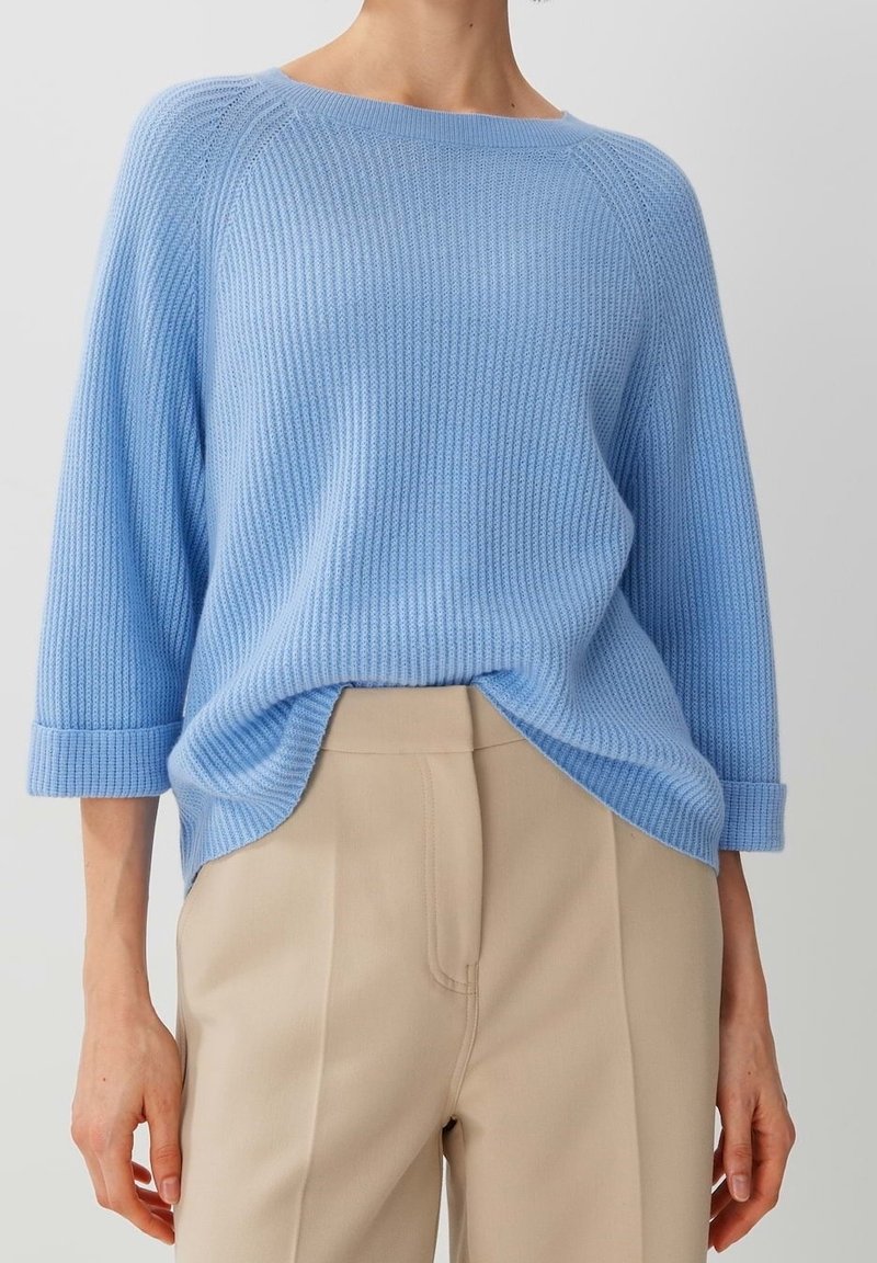 Pull léger bleu à côtes avec des manches trois-quarts, offrant une coupe décontractée et de légentes fentes sur les côtés à l'ourlet, associé à un pantalon beige.