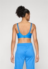 Blå idretts-BH med justerbare stropper som har "UNDER ARMOUR"-logo; glatt stoff med myk tekstur, kombinert med matchende blål øye joggebukser.