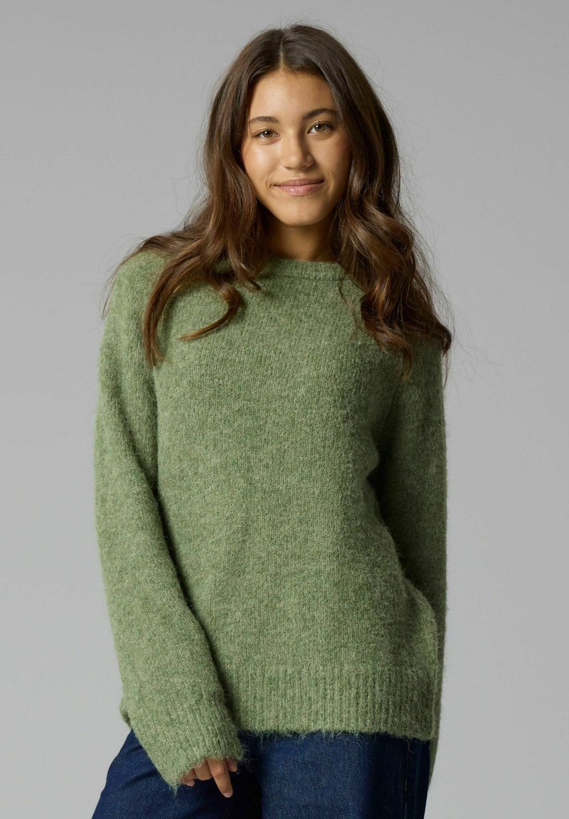 Jeune femme aux longs cheveux bruns portant un pull vert duveteux et un jean bleu, debout devant un fond gris uni.