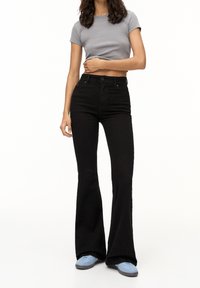 Jean noir évasé en denim, taille haute, associé à un t-shirt gris court et des baskets bleues. Texture lisse, design simple.