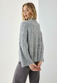 Suéter gris de punto con patrón de trenzas, mangas anchas y dobladillo acanalado. Textura suave y esponjosa, usado sobre pantalones grises, mostrado desde atrás.