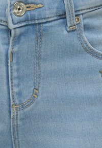 Lichtblauwe denimstof met gouden stiksels en een metalen knoopsluiting. Bevat een voorzak en subtiele slijtage details.