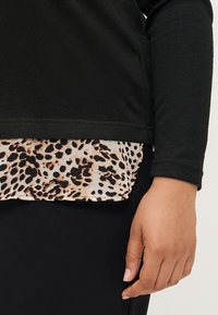 Černý top s dlouhým rukávem, který má texturovanou látku překrývající kontrastní leopardí vzor na spodním lemu. Detail rukávu a ruky.