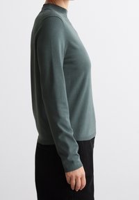 Grön långärmad tröja med hög krage, åtsittande design, mjuk textur och ribbade cuffs, i kombination med svarta byxor.