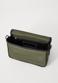 Borsa in nylon verde oliva con cerniera nera, apertura superiore e interno liscio. Presenta un portacarta con logo all'interno e bordi resistenti.