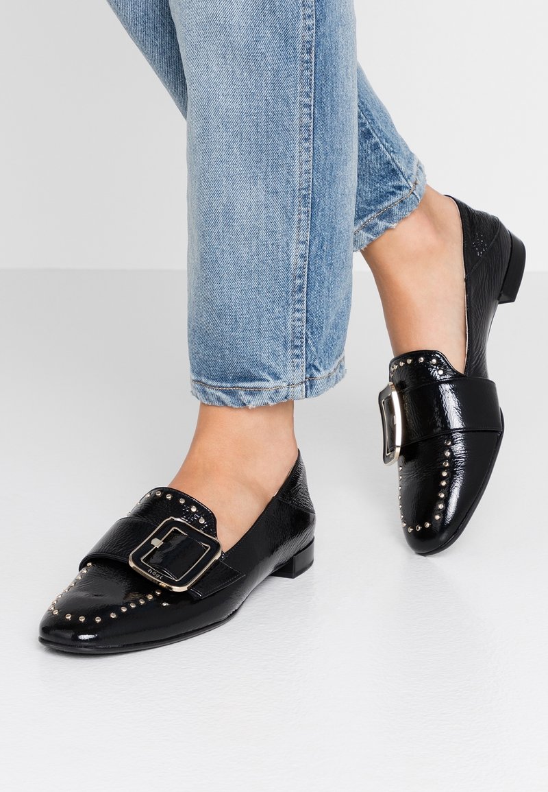 Mocassins en cuir noir avec un bout rond, une boucle décorative, des accents cloutés le long du capuchon, et un talon bas. Associés à un jean bleu retroussé.