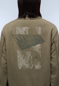 Olivengrøn sweatshirt med et struktureret print, der viser teksten "NAPAPIJRI" i mørkegrøn og en grafik fra 1987. Blødt stof med en afslappet pasform.