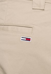 Béžové bavlněné kalhoty mají zadní kapsu s malým vyšívaným logem v červené, bílé a modré barvě, které dodává designu jedinečný detail.