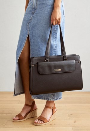 Mujer sosteniendo un gran bolso de cuero negro, con una falda de mezclilla azul clara con una abertura frontal y sandalias con cuña marrones.