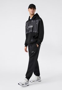 Sudadera negra con gráficos de "Champion" y "FASHION"; combinada con joggers negros con detalle de logo. Zapatillas deportivas blancas completan el conjunto.