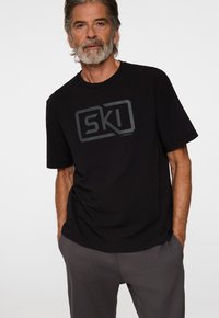 Mies, jolla on harmaat hiukset ja parta, pukeutuneena mustaan t-paitaan, jossa on "SKI" -grafiikka, ja harmaisiin housuihin, seisoo yksinkertaista valkoista taustaa vasten.