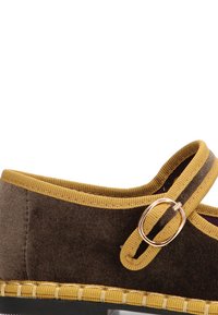 Zapato de tela marrón con ribete amarillo, con un acento metálico redondo y costuras a lo largo de la suela. Correa ajustable para el cierre.