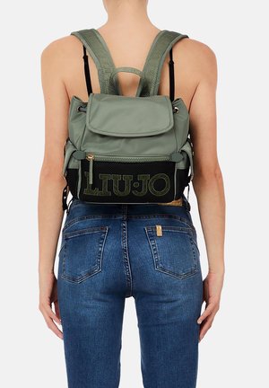 Persona che indossa jeans blu e uno zaino verde Liu Jo con spallacci imbottiti, vista da dietro contro uno sfondo semplice.