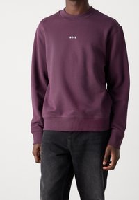 Fialová mikina s kulatým výstřihem, žebrovanými manžetami a lemem, s nápisem "BOSS" bílou barvou přes hrudník. Nošena s černými džínami.