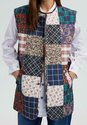Kobieta nosząca kamizelkę bez rękawów typu patchwork z różnymi wzorami kwiatowymi, w kratkę i paisley, na białej koszuli zapinanej na guziki i dżinsach.
