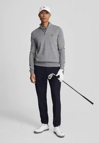 Siv pulover na zadrgo z majhnim rumenim logotipom, mornarsko modre hlače, beli supergi in bela kapa; drži golf palico z roko v rokavici.