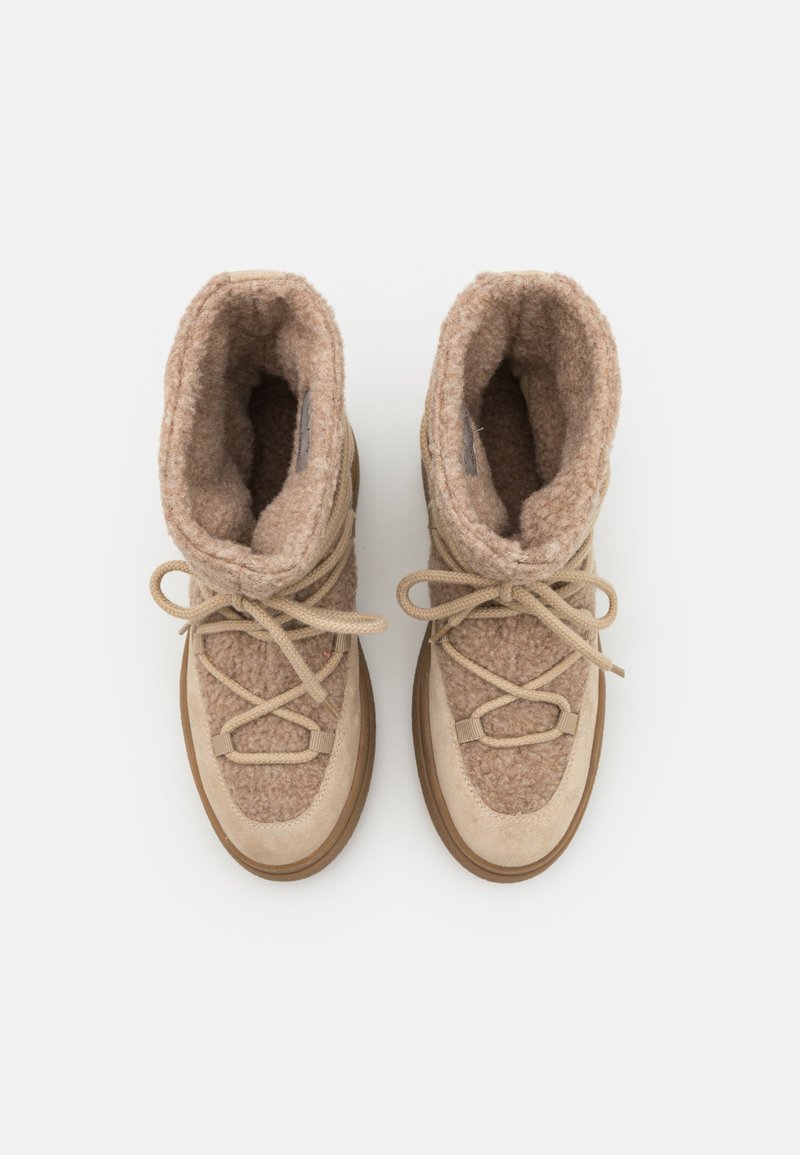 Ein Paar beiger, flauschiger Winterstiefel mit Schnürsenkeln, dickem, weichem Futter und Gummisohlen, von oben auf einem weißen Hintergrund gezeigt.