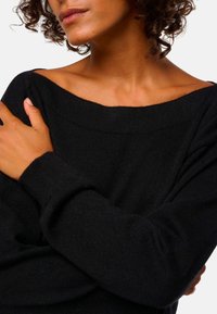 Frau mit mittlerem Hautton und lockigem Haar, trägt einen schwarzen schulterfreien Pullover, die Arme über der Brust verschränkt, vor einem schlichten Hintergrund.