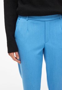 Heledad sinised kohandatud püksid, mis on valmistatud tekstuurilisest kangast, küljetaskutega ja lameda eesmiuga, sobitatud musta kootud sviitriga.