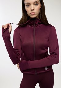 Giacca sportiva bordeaux con zip frontale, aperture per il pollice nelle maniche e colletto alto. Tessuto liscio e elasticizzato; logo sul bordo inferiore.