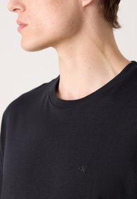 Ung person iført en ensfarget svart t-skjorte med rund hals og liten svart "cK" logo på brystet, rettet mot venstre.