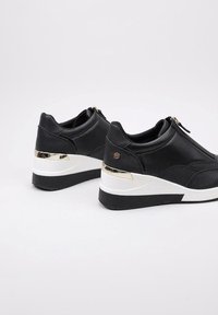 Zapatillas negras con un acabado texturizado, entresuela blanca, detalles dorados y cierre de cremallera. Tacón ligeramente elevado para mayor altura.