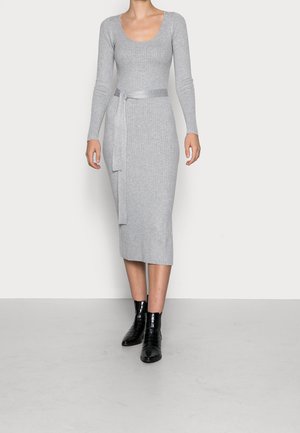 Kobieta ubrana w dopasowaną, prążkowaną, długorękawową szarą sukienkę midi z paskiem w talii oraz czarne botki na prostym tle.