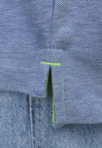 Blue textured fabric with a green accent stitch at the hem, partially covering denim pants, showcasing a split detail at the edge.