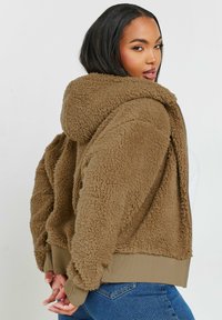 Brun, fluffig huvtröja med dragkedja framtill, ribbat nederkant och ärmar. Mjuk textur och avslappnad passform med en stor huva.