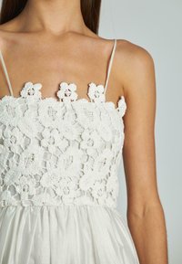 Witte jurk met een bloemen kant lichaam en ingewikkelde haakdetails, met dunne bandjes die leiden naar een vloeiende, soepele rok.