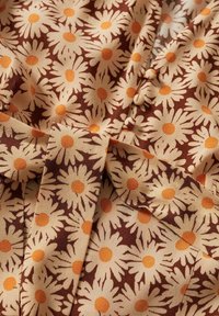 Tela floral que presenta margaritas blancas con centros naranjas sobre un fondo marrón. Textura suave con un leve brillo; incluye detalles de botones.